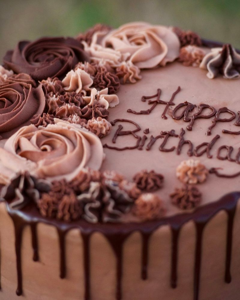Üstünde krema gülleri ve çikolatayla yazılmış Happy Birthday yazısı olan çikolatalı pasta